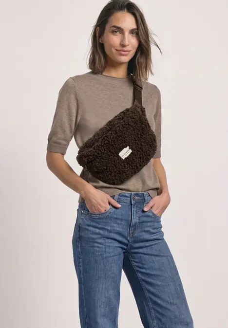Eine Frau trägt einen beigen Kurzarm-Pullover, blaue Jeans und eine braune, flauschige Bauchtasche vor neutralem Hintergrund.