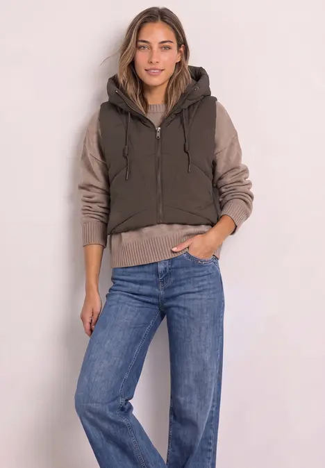 Frau in dunkelgrüner Steppweste mit Kapuze, beigem Pullover und blauen Jeans vor neutralem Hintergrund.