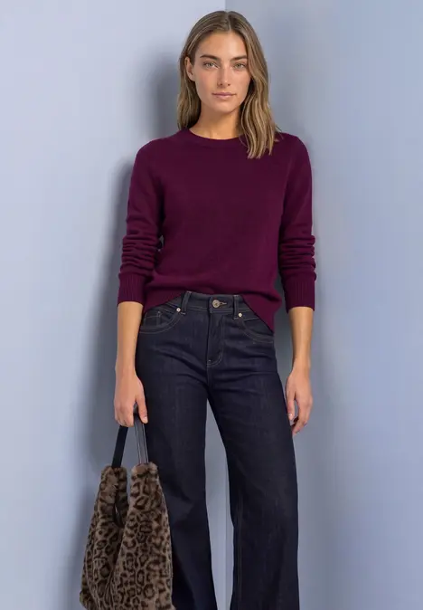 Frau in dunkelviolettem Pullover, weiten Jeans und Leoparden-Tasche vor hellem Hintergrund.