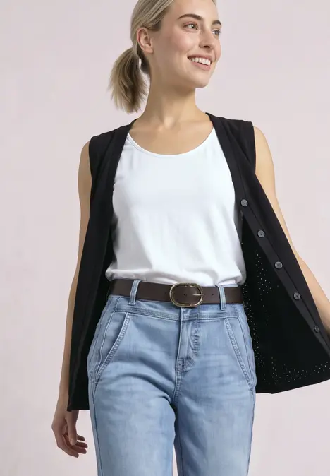 Lächelnde Frau mit blondem Haar, weißem Tanktop, schwarzer Weste offen, hellblauen Jeans und Gürtel.