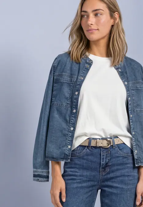 Frau in weißem T-Shirt und Jeans mit lässiger Jeansjacke vor neutralem Hintergrund.