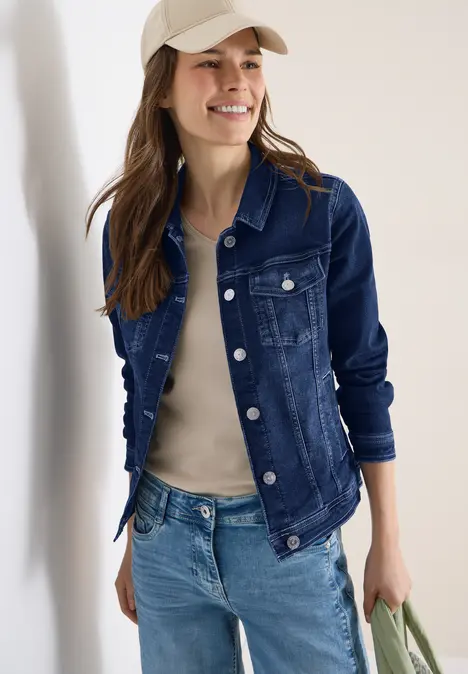 Person trägt Jeansjacke, helles Top, Jeans und hält eine grüne Tasche.