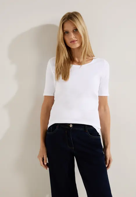 Eine blonde Frau mit weißem T-Shirt und blauer Jeans steht vor einer neutralen Wand und blickt in die Kamera.