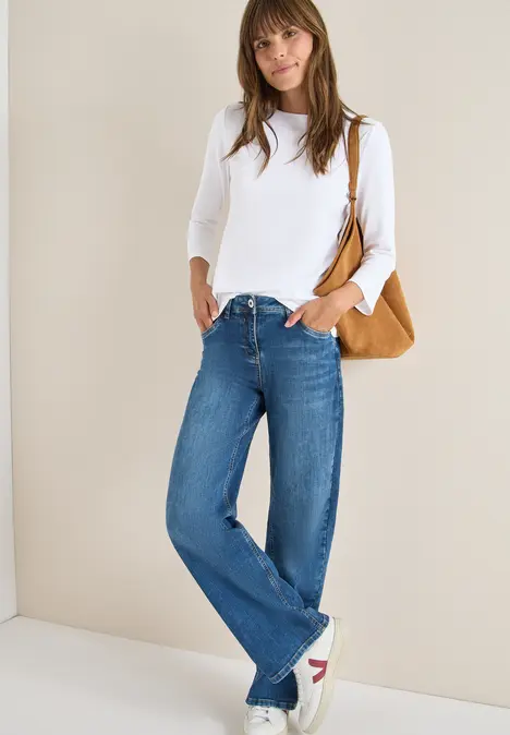 Frau in weißem Langarmshirt und weiten Jeans, weiße Sneakers, braune Tasche, vor neutralem Hintergrund.