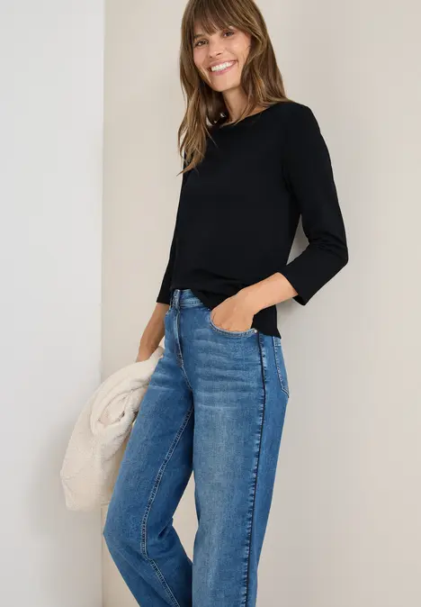 Fröhliche Frau in schwarzem Langarmshirt und blauen Jeans, hält eine beige Jacke in der Hand.