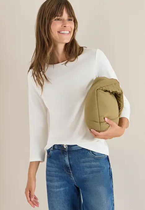 Frau in weißem Langarmshirt und Jeans lächelt und hält eine olivgrüne Steppjacke.