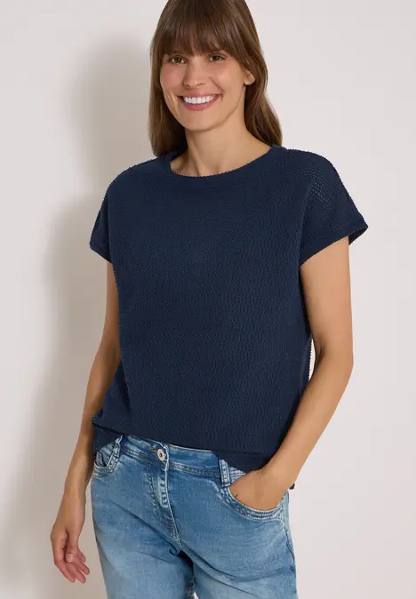 Frau mit dunkelblauem kurzärmligem Oberteil und Jeans, lächelnd mit den Händen in den Taschen.