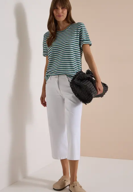 Frau trägt gestreiftes T-Shirt, weiße Culotte, beige Mules und hält eine schwarze Felltasche.