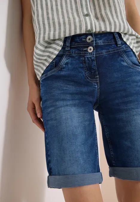 Frau in gestreiftem Top und hochgekrempelter Jeans-Shorts vor weißer Wand