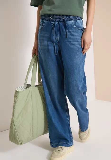 Eine Person trägt weite blaue Jeans, weiße Sneakers und hält eine gesteppte grüne Tasche.