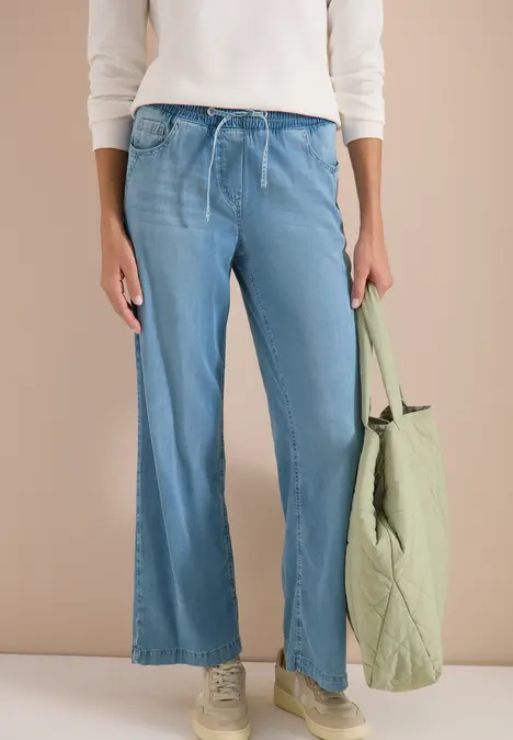 Frau in hellblauen, weitem Jeanshosen mit Kordelzug, weißem Oberteil, grünem Stoffbeutel und beigen Sneakers.