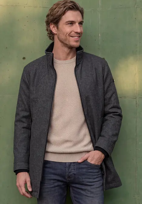 Ein Mann trägt grauen Mantel, beiges Shirt und Jeans, lächelt vor grünem Hintergrund.