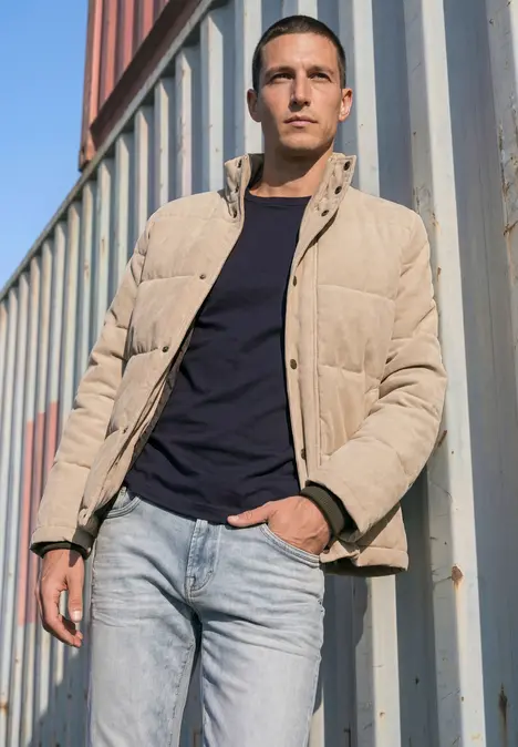 Ein ernst blickender Mann in beiger Pufferjacke und Jeans lehnt an Containern.