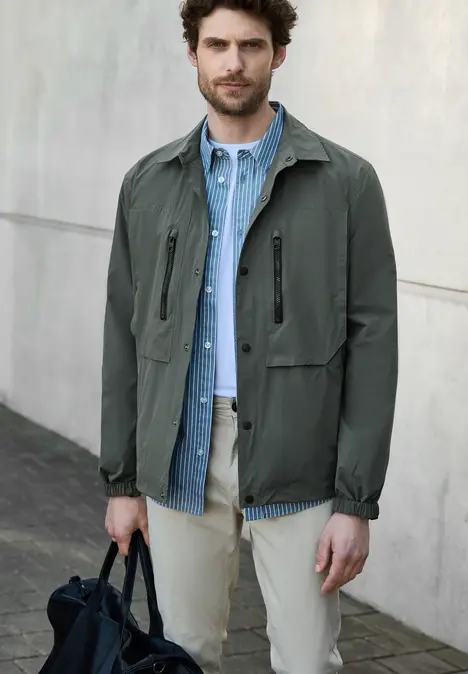 Homme en veste verte, chemise bleue et sac noir, debout dehors.