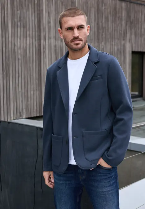 Man in spijkerbroek, wit t-shirt en donkerblauw jasje voor houten wand.