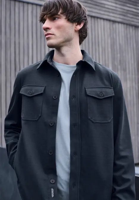 Jeune homme en chemise overshirt noire et t-shirt blanc appuyé devant un panneau en bois.
