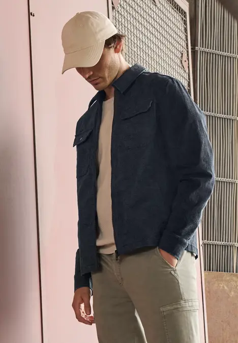 Homme en veste en jean, T-shirt beige, pantalon et bob beige devant un mur rose et un portail metal.