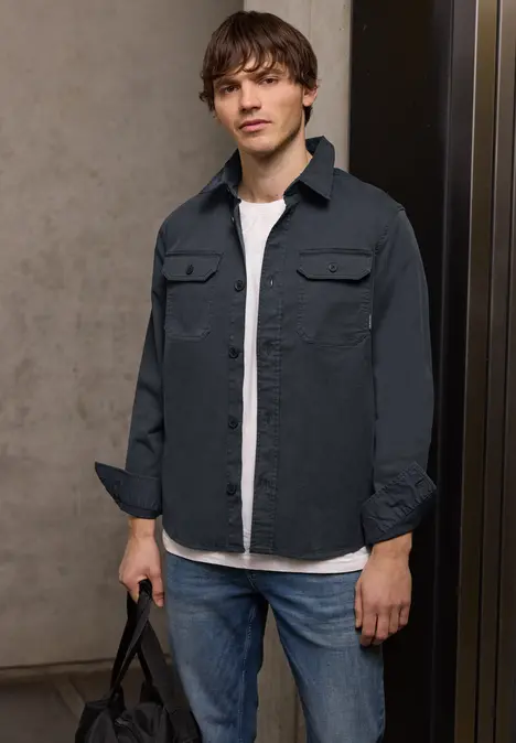 Un homme en jean, t-shirt blanc et chemise gris foncé se tient avec un sac à la main.