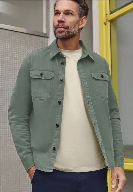 Un homme pose décontracté en surchemise verte et T-shirt blanc devant un cadre de porte jaune.