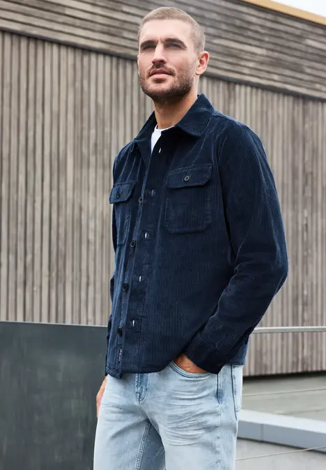 Mann mit kurzem Haar in dunkelblauer Cordjacke und hellen Jeans vor Holzwand.