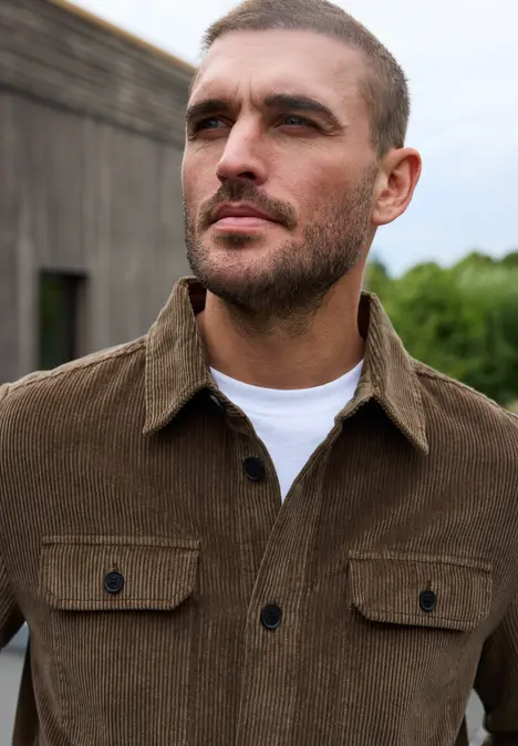 Ein Mann mit kurzem Haar und Bart trägt eine braune Cordjacke vor einem Holzhintergrund.