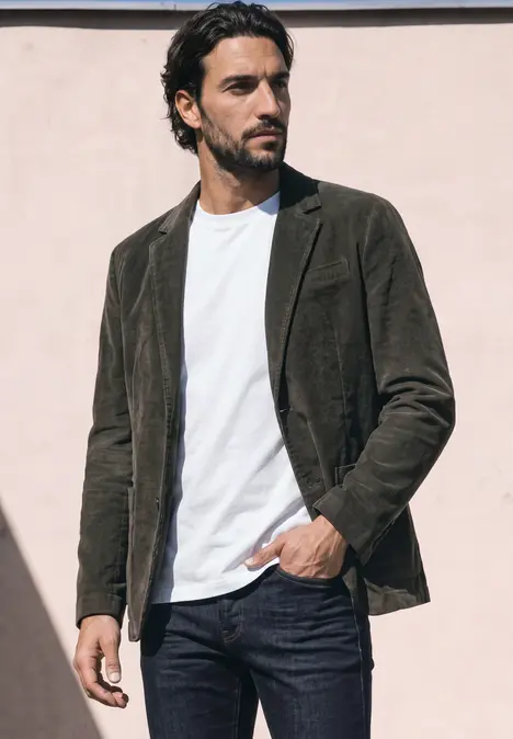 Ein Mann steht vor einer hellen Wand in weissem T-Shirt, braunem Cordblazer und dunkler Jeans.