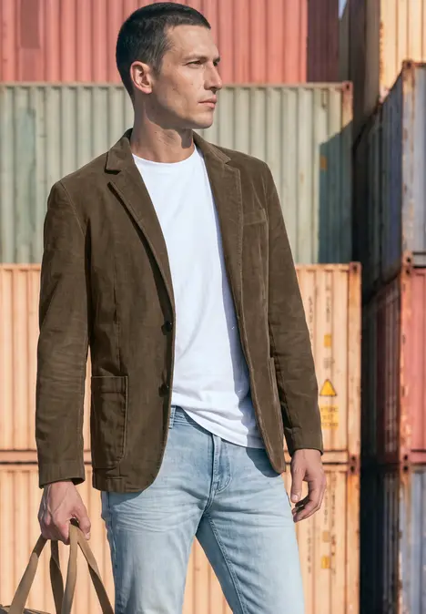 Mann in weißem T-Shirt, brauner Jacke und Jeans steht vor gestapelten Frachtcontainern.