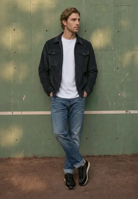 Un jeune homme en jean, t-shirt blanc et veste noire pose devant un mur vert.