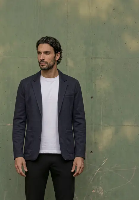 Homme devant un mur vert en blazer bleu marine, t-shirt blanc et pantalon noir.