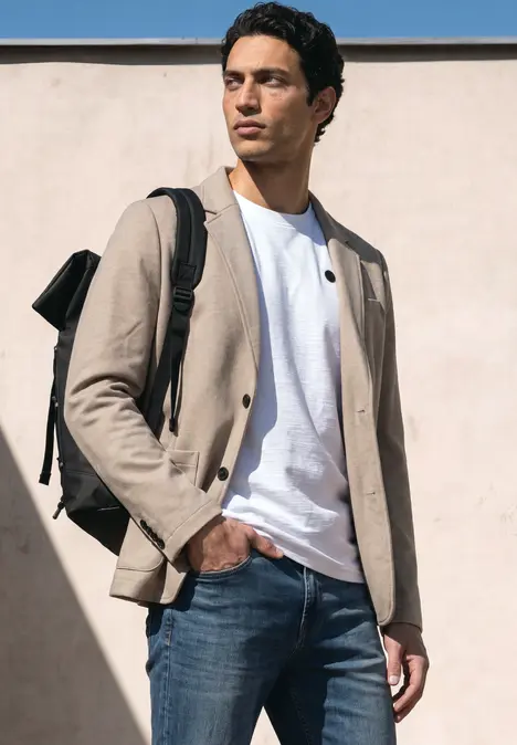 Un homme se tient devant un mur, vêtu d'une veste beige, d'un T-shirt blanc, d'un jean bleu et d'un sac à dos.