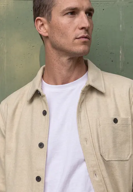Nahaufnahme eines jungen Mannes mit kurzem Haar, der ein beiges Hemd und ein weißes T-Shirt trägt.