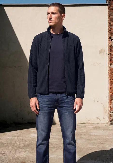 Ein Mann in Jeans, T-Shirt und Jacke steht vor einer hellen Wand im Freien.