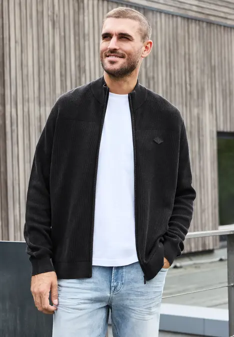 Ein lächelnder Mann in schwarzer Jacke, weißem T-Shirt und Jeans lehnt entspannt an einem Geländer.