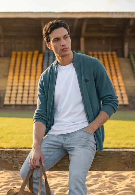 Ein junger Mann in Jeans und Jacke lehnt mit Tasche vor leeren Tribünen auf einem Sportplatz.
