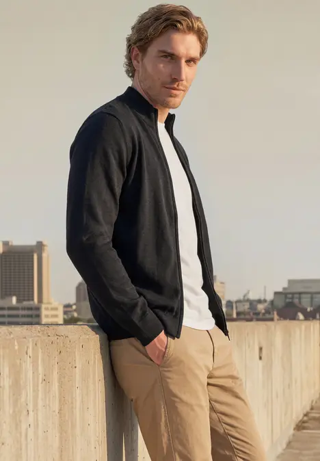 Ein Mann in schwarzer Jacke, weißem T-Shirt und beiger Hose lehnt entspannt an einer Dachbrüstung mit Stadtblick.