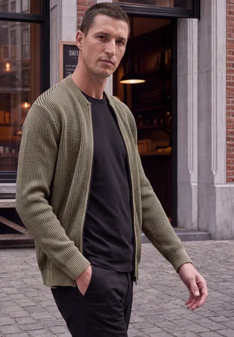 Ein Mann in grüner Strickjacke und schwarzem T-Shirt läuft vor einem Café in der Stadt.
