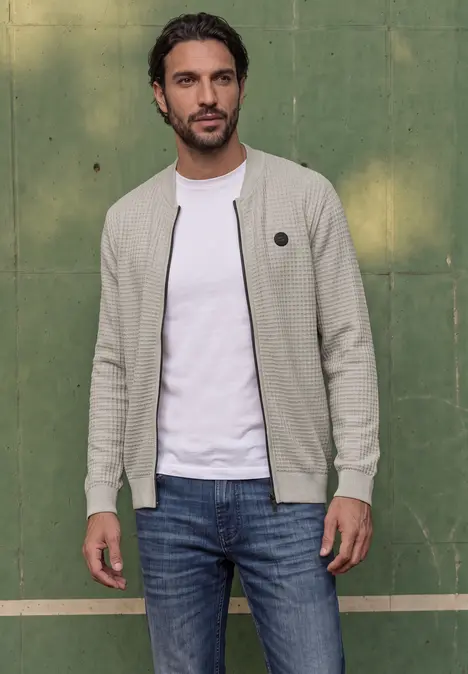 Ein Mann in heller Strickjacke und Jeans steht lässig vor einer grünen Wand.