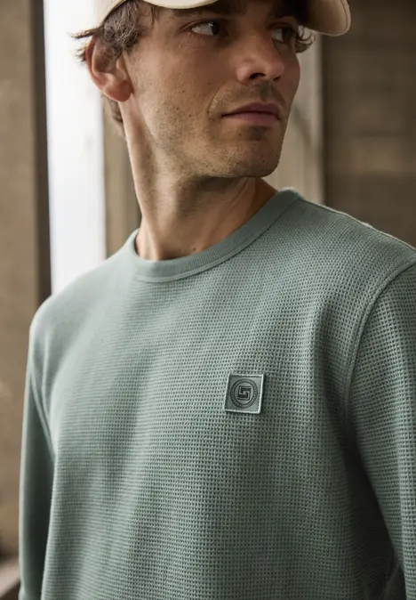 Homme portant un pull vert texturé, debout dans une pièce lumineuse devant un mur en briques.