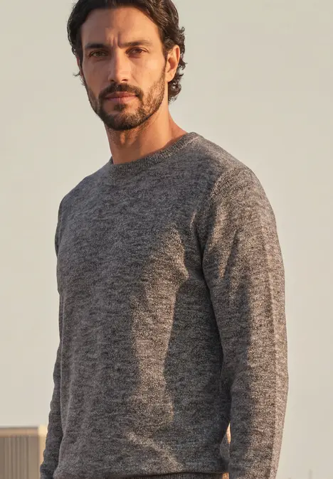 Ein dunkelhaariger Mann mit Drei-Tage-Bart trägt einen grauen Pullover vor neutralem Hintergrund.