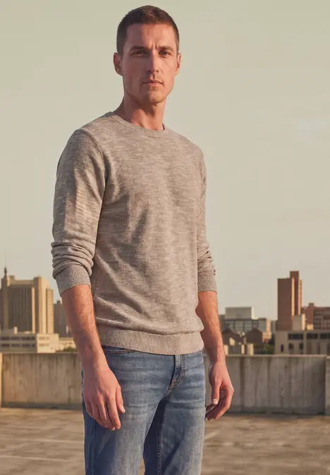 Ein Mann in grauem Pullover und Jeans steht auf einem Dach mit Stadtgebäude im Hintergrund.