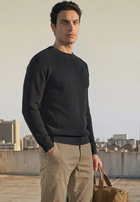 Ein Mann in schwarzem Pullover und hellen Hosen steht auf einem Dach und hält eine Tasche in der Hand.