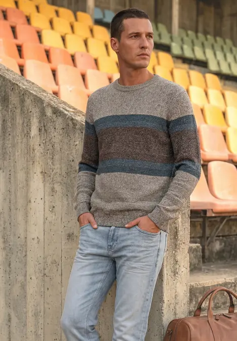 Ein Mann lehnt an einer Stadiontribüne, trägt gestreiften Pullover, Jeans und eine braune Tasche.