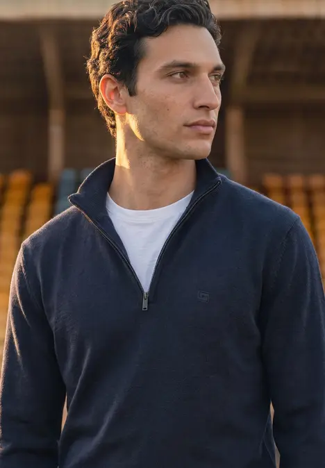 Ein Mann in Viertel-Zip-Pullover und weißem T-Shirt vor Tribünen im warmen Abendllicht.