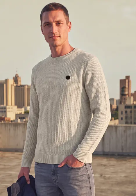 Ein Mann in einem hellen Pullover steht auf einem Dach, im Hintergrund die Stadt.