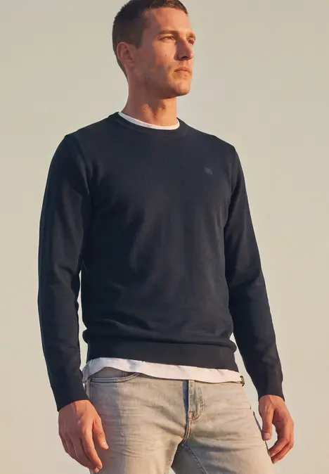 Ein Mann steht im Freien und trägt einen dunklen Pullover, ein weißes T-Shirt und helle Jeans.