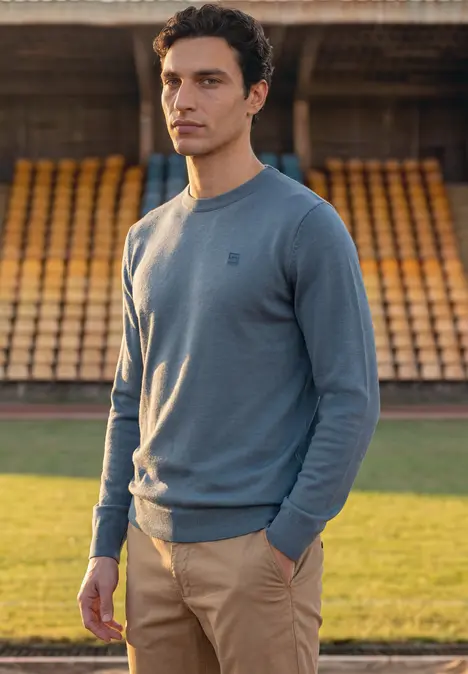 Homme en pull bleu clair et pantalon beige devant des gradins vides.