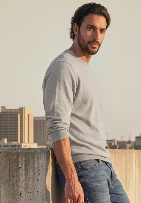 Ein lässig gekleideter Mann lehnt sich vor einer Betonmauer ab, trägt grauen Pullover und Jeans.