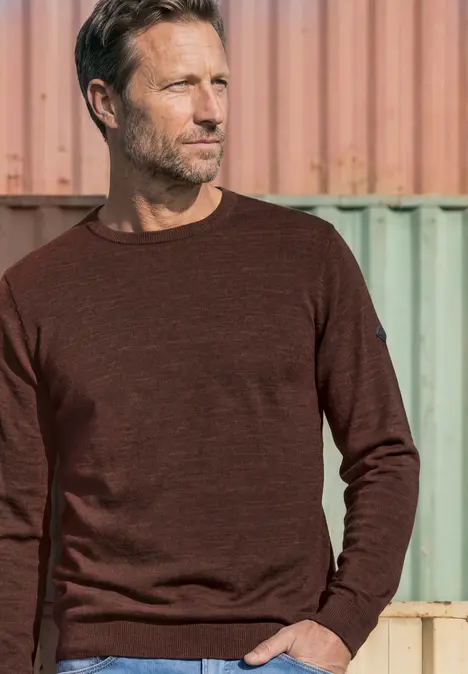 Ein Mann in braunem Pullover steht vor Containern und blickt zur Seite.