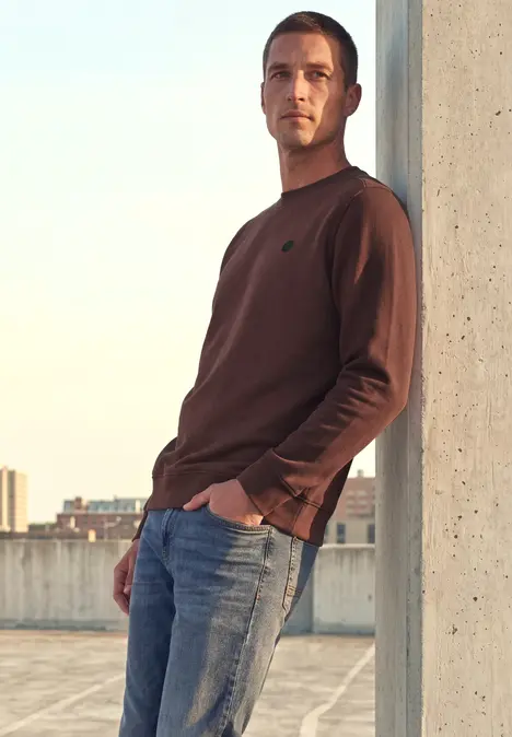 Ein lässig gekleideter Mann lehnt an einem Pfeiler auf einem Parkdeck, die Stadt im Hintergrund.