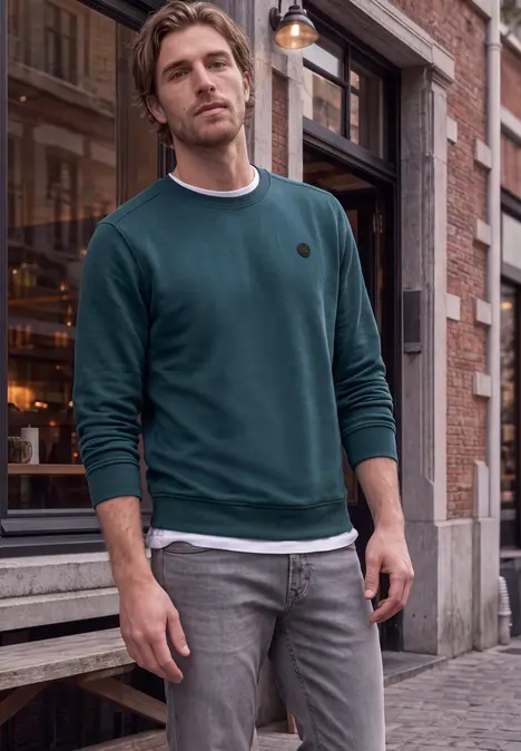 Ein Mann in grünem Pullover und grauen Jeans steht entspannt vor einem Laden auf Kopfsteinpflaster.